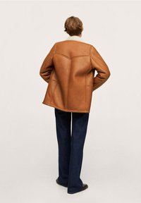 Persona vista da dietro che indossa una giacca shearling marrone con colletto bianco, jeans blu scuro e scarpe nere, in piedi su uno sfondo beige.