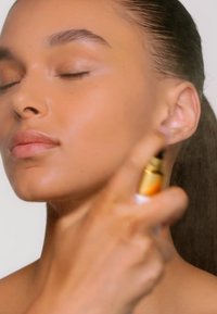 Flacone spray per vaporizzatore viso con dettaglio dorato, tenuto in mano. Lo sfondo è neutro. Il soggetto ha gli occhi chiusi e una carnagione liscia e luminosa.