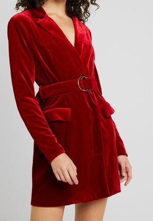 Femme portant une robe blazer en velours rouge ceinturée avec poches à rabat et manches longues sur fond gris.
