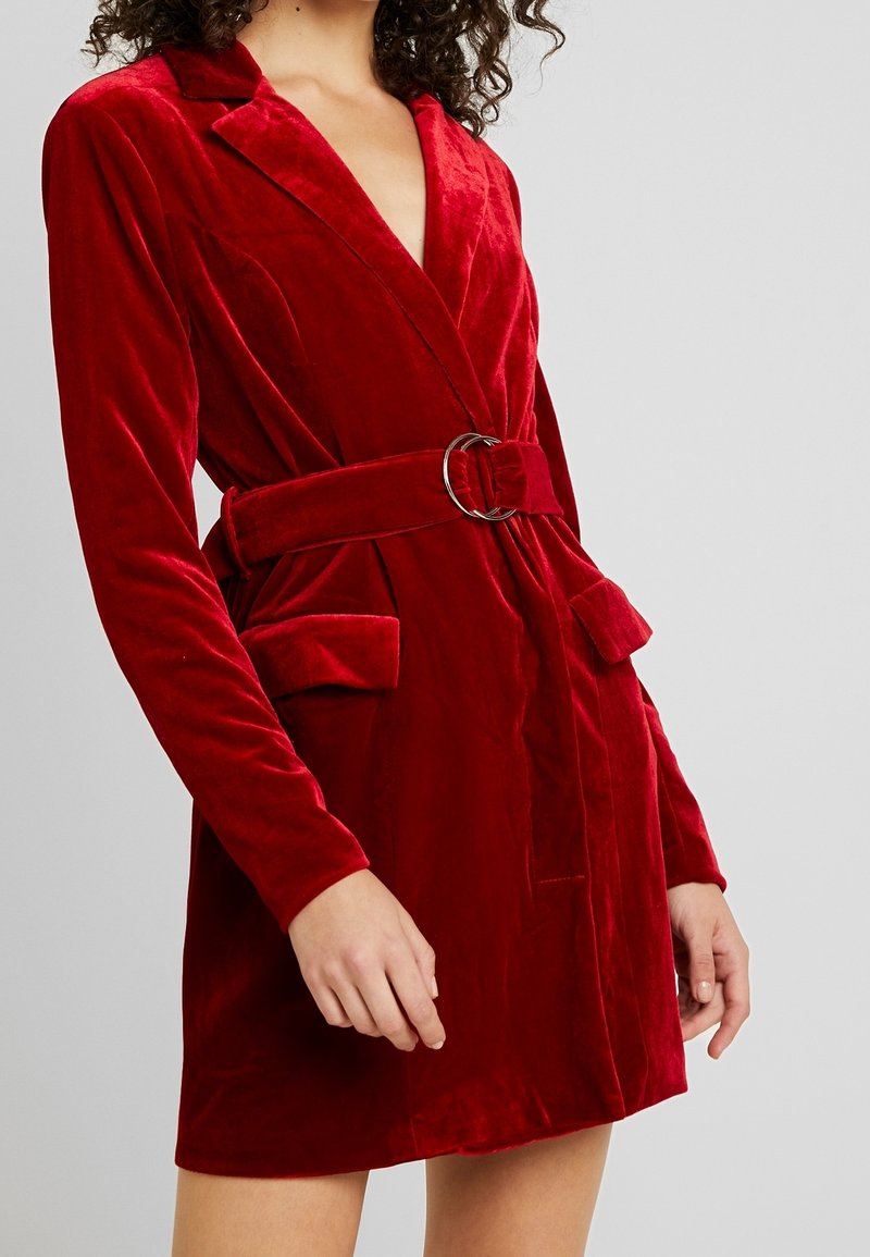 Femme portant une robe blazer en velours rouge ceinturée avec poches à rabat et manches longues sur fond gris.