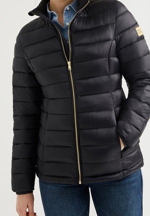 Schwarze gesteppte Jacke mit goldenem Reißverschluss, getragen über einem blauen Hemd mit Knöpfen und blauer Jeans, rechte Hand in der Tasche.