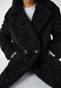 Manteau noir en peluche avec un matériau texturé et duveteux. Comprend un grand col, des boutons croisés et des poches latérales. Porté par-dessus un pull gris clair.