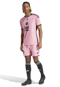 Maillot de football rose avec des accents noirs, arborant un logo et un motif de couronne. Accompagné de shorts assortis et de chaussettes noires montantes à rayures.