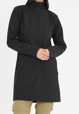 Kvinde iført en lang, sort lynlåsjakke med hætte, hænderne foldet, kombineret med beige bukser mod en ensfarvet baggrund.