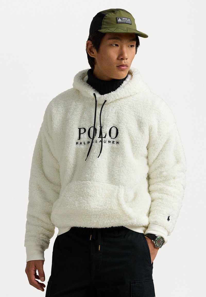 Hvit, fluffy hettegenser med svart "POLO RALPH LAUREN" logo, snor og sidelommer. Kombinert med en grønn caps og svarte bukser.