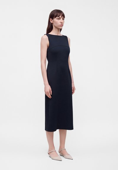 Femme portant une robe noire sans manches, longueur aux genoux, avec un décolleté carré et des ballerines beiges pointues, debout sur un fond blanc uni.