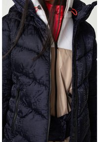 Námořnicky modrá prošívaná péřová bunda s kapucí, kontrastními zipy a výrazným vzorem loga. Pod ní vrstvy v béžové a červené barvě.