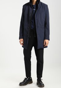 Cappotto blu navy con colletto alto, chiusura a bottone e tessuto strutturato, indossato sopra una camicia scura e pantaloni neri con calzini a pois e scarpe.