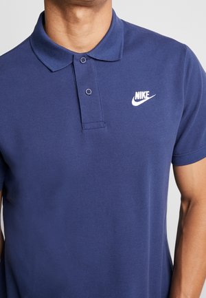 Man draagt een donkerblauw Nike polohemd met twee knoopsluiting en een wit Nike-logo op de linkerborst.