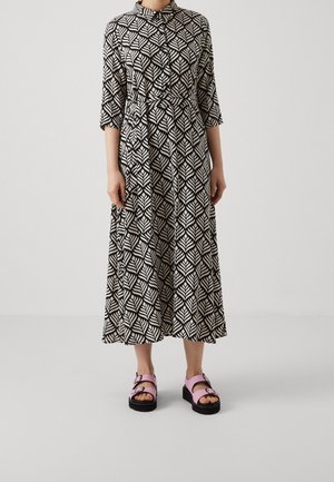Personne portant une longue robe à motifs noir et blanc avec une ceinture à la taille et des manches trois-quarts, associée à des sandales à plateforme roses.