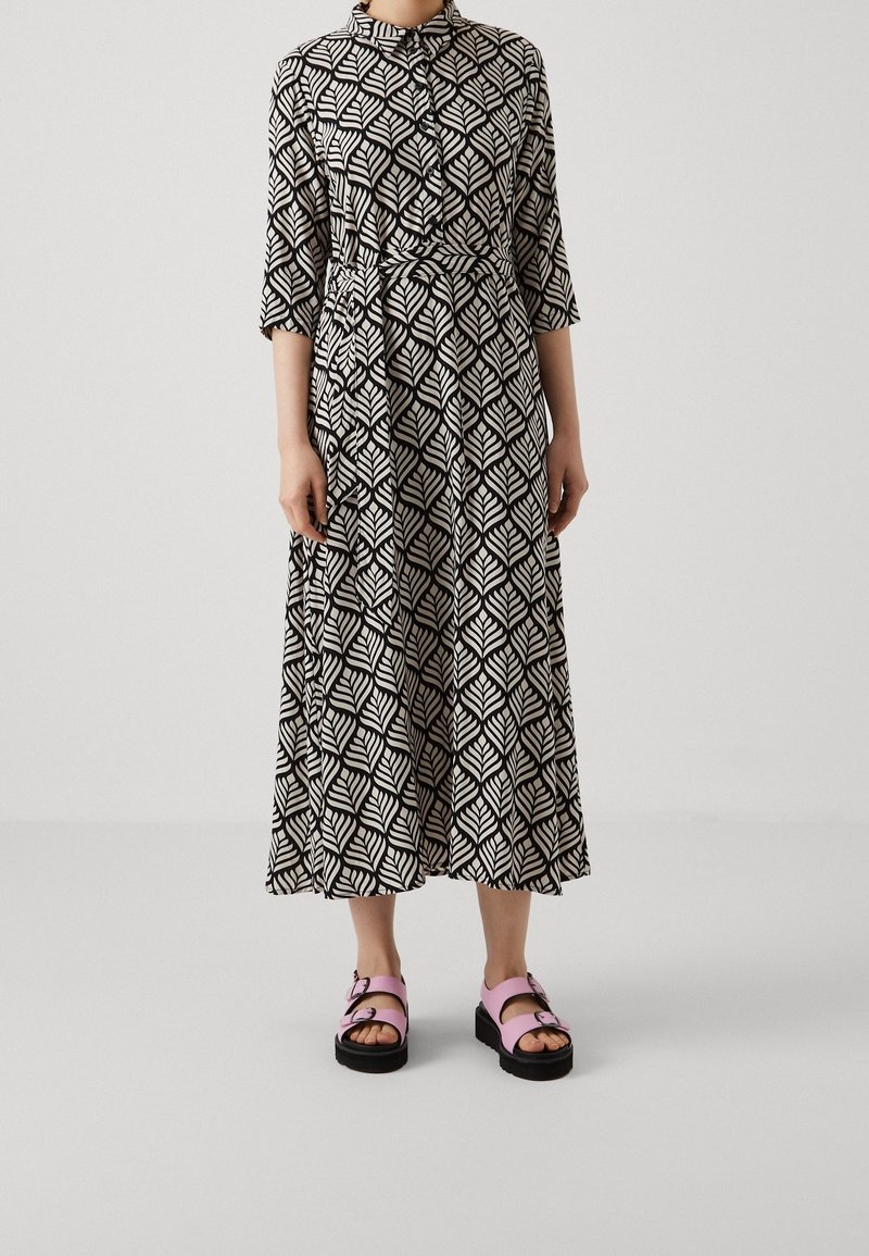 Personne portant une longue robe à motifs noir et blanc avec une ceinture à la taille et des manches trois-quarts, associée à des sandales à plateforme roses.