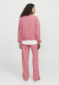 Roza bombažna majica s kapuco z spušenimi rameni in rebrastimi manšetami, v kombinaciji z ujemajočimi se ohlapnimi hlačami. Gladka tekstura, minimalistična zasnova.