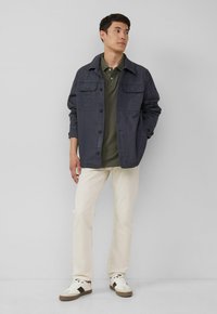 Marineblaues Baumwolljackett mit zwei vorderen Taschen, grünem Polohemd darunter, cremefarbenen geraden Hosen und weißen Sneakers mit braunen Akzenten.