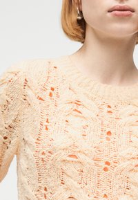 Κοντινό πλάνο ανοιχτόχρωμου ροδακινί πλεκτού πουλόβερ με περίπλοκα σχέδια κοτσίδας και δαντέλας στο άνω μέρος του κορμού και του λαιμού ενός ατόμου.