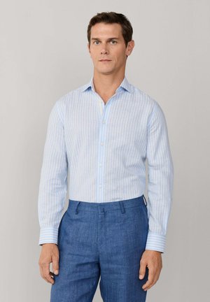 Mann trägt ein hellblau gestreiftes langärmeliges Hemd, in blaue Hose gesteckt, steht vor einem einfachen hellen Hintergrund.