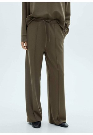 Pantalones marrones holgados con costuras delanteras, cintura con cordón, usados con top a juego y zapatos negros, de pie frente a un fondo liso.