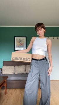 Kartonová krabice Zalando držena osobou, která má na sobě bílý crop top a šedé široké kalhoty. Místnost má zelené stěny a sedačku.