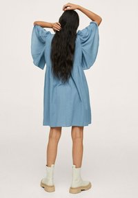 Robe à carreaux bleu clair avec des manches larges, un dos froncé et une coupe loose, associée à des bottes de cheville à plateforme crème, vue de dos.