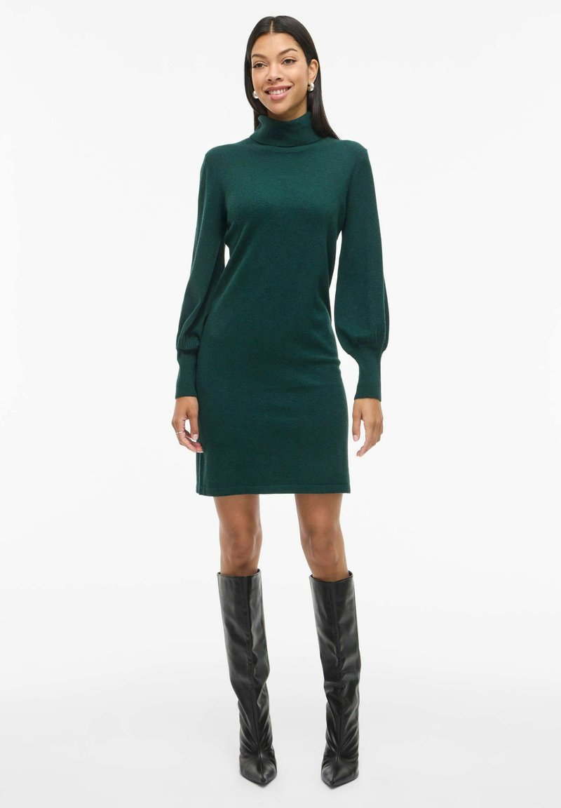Vestido de punto verde con un escote alto y mangas largas abullonadas, combinado con botas negras hasta la rodilla. Textura suave y diseño ajustado.