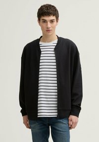 Felpa nera con zip e colletto rotondo. Indossata sopra una t-shirt a righe bianche e blu navy. Presenta polsini a coste e una vestibilità comoda.