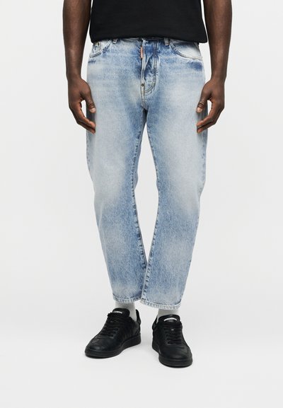 Homme portant un jean bleu clair délavé et des baskets noires, debout devant un fond blanc uni.