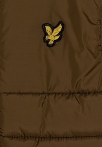 Giubbotto puffo marrone con texture trapuntata e logo di un uccello giallo ricamato. Il tessuto presenta lucentezza e una finitura liscia.