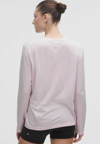 Cămașă cu mânecă lungă dintr-un material roz deschis, cu un model texturat; are un guler rotund și un logo pe spate. Poartă cu pantaloni scurți negri.