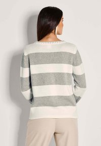 Frau mit langen dunklen Haaren, die große Creolen trägt, in einem hellgrau-weiß gestreiften Pullover und beigen Hosen, die von der Kamera abgewandt vor einem schlichten Hintergrund steht.