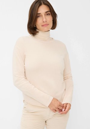 Beige turtleneck sweater with long sleeves, ribbed texture, and a slightly relaxed fit, paired with light-colored pants.