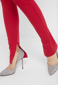 Červené pouzdrové kalhoty se bočními rozparky v kombinaci se stříbrnými třpytivými vysokými podpatky. Boty mají špičaté špičky a stiletto podpatky.