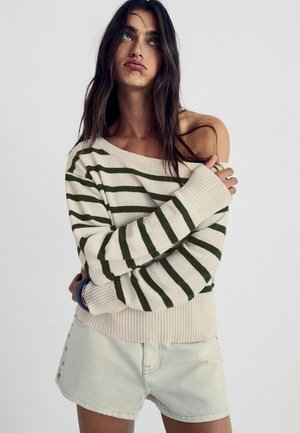 Žena s dlhými tmavými vlasmi nosí béžový sveter bez ramienok so zelenými pruhmi a svetlé šortky, pózuje so skríženými rukami na bielom pozadí.