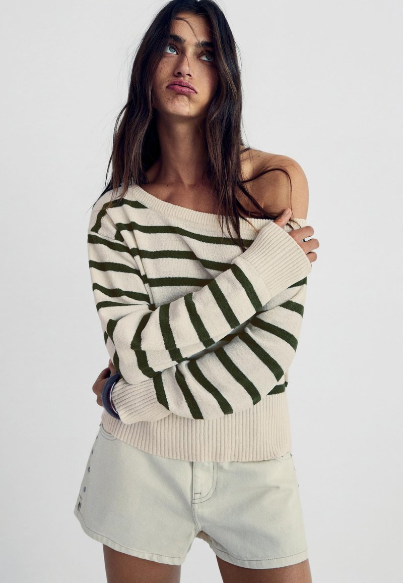 Femme aux longs cheveux foncés portant un pull beige à rayures vertes, décolleté épaules, et un short clair, posant les bras croisés devant un fond blanc.