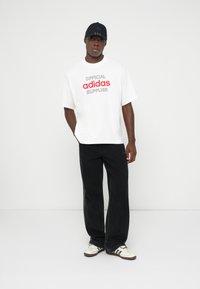 Moški oblečen v belo majico Adidas z rdečim napisom, široke črne jeans hlače, bele superge in črno kapo, stoji pred enobarvno belo ozadje.
