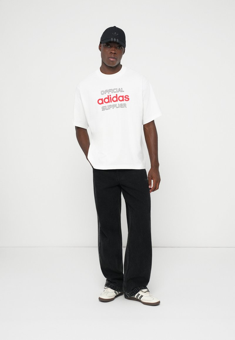 Hombre con camiseta blanca de Adidas con texto rojo, pantalones anchos negros, zapatillas blancas y gorra negra, de pie contra un fondo blanco liso.