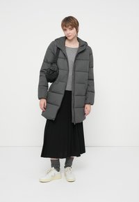 Kvinde iført en grå polstret jakke, grå sweater, sort plisseret nederdel, grå sokker, hvide sneakers og en lille sort skuldertaske på hvid baggrund.