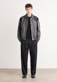 Zwarte leren varsityjack met grijze accenten, drukknoopsluiting en een logo patch. Draagt over een zwarte sweater en gecombineerd met losse zwarte pants.