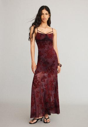 Femme aux longs cheveux foncés portant une robe maxi florale rouge foncé transparente sans manches et des sandales noires à talons, debout devant un fond uni.
