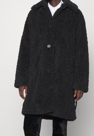 Person wearing long black textured teddy coat with oversized collar and button closure over white shirt and black pants.