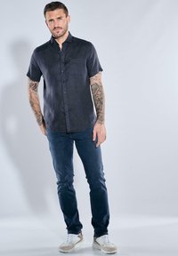 Kurzärmliges, dunkelblaues Hemd mit Knöpfen und einer Brusttasche. Kombiniert mit schmal geschnittenen, dunklen Jeans und hellen Sportschuhen.