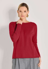 Eine Frau mit glatt nach hinten gekämmtem Haar, die große goldene Creolen, einen roten Langarmpullover und graue, gefaltete Hosen trägt, posiert vor einer hellen Wand.