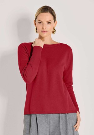 Eine Frau mit glatt nach hinten gekämmtem Haar, die große goldene Creolen, einen roten Langarmpullover und graue, gefaltete Hosen trägt, posiert vor einer hellen Wand.