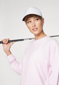 Ροζ, υφασμάτινο πουλόβερ με διαμαντένιο μοτίβο και ραμμένο μανίκι; λευκό καπέλο; κρατάει μια γκολφ κλαμπ στον ώμο; ουδέτερο φόντο.