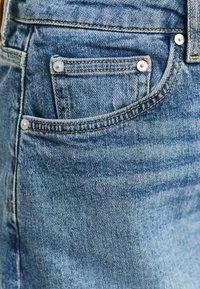 Lichtblauwe denim jeans hebben een voorzak met zilverkleurige hardware en contrasterend gele stiksels, en tonen een versleten textuur en een klassieke snit.