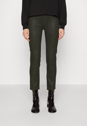 Kobieta ubrana w ciemnozielone, dopasowane spodnie oraz czarne, błyszczące botki, stojąca na jednolitym, jasnym tle.