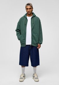 Groene zip-up hoodie met trekkoord, twee voorzakken; witte t-shirt eronder, gecombineerd met losse blauwe denim shorts en witte sneakers.