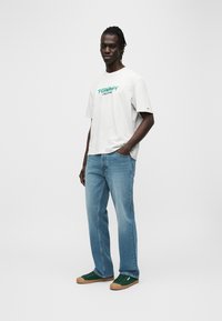 Γκρι βαμβακερό t-shirt με πράσινο λογότυπο "TOMMY JEANS", συνδυασμένο με ανοιχτόχρωμο μπλε τζιν παντελόνι και πράσινα αθλητικά παπούτσια με σόλες από καουτσούκ. Απλά στοιχεία σχεδίασης.