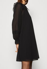 Vestido de gasa negro con mangas largas recogidas, escote con volantes y detalle de lazo. Presenta una silueta suelta y fluida, además de una textura transparente.