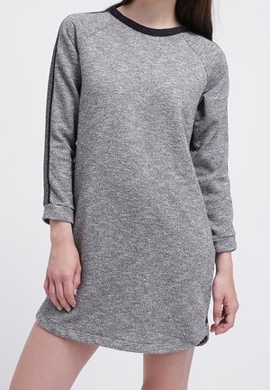 Femme portant une robe-pull à manches longues en maille grise avec des rayures noires sur les manches, debout devant un fond uni.