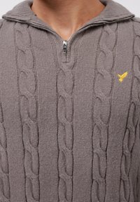 Hellbrauner Pullover aus Kabelstrick mit einem Halb-Reißverschluss am Hals und einem kleinen gelben, gestickten Vogel-Logo auf der Brust.