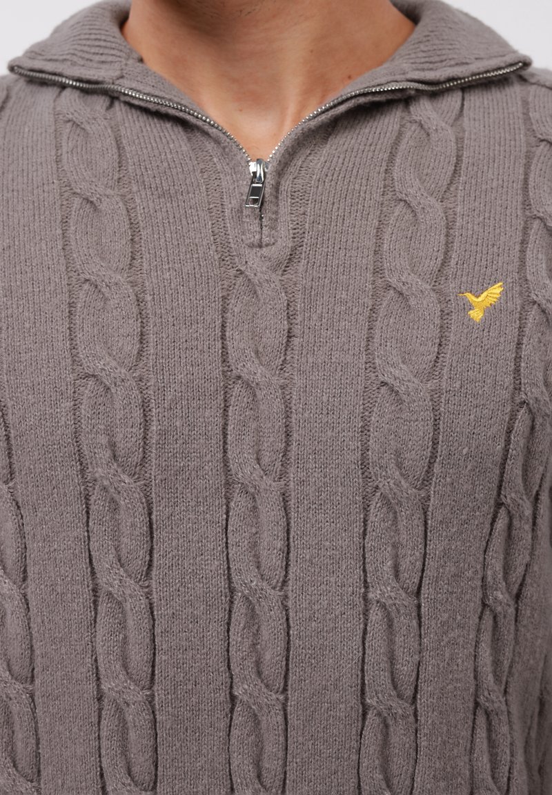 Jasnobrązowy sweter w splot z półsuwnym kołnierzykiem i małym żółtym haftowanym logo ptaka na piersi.